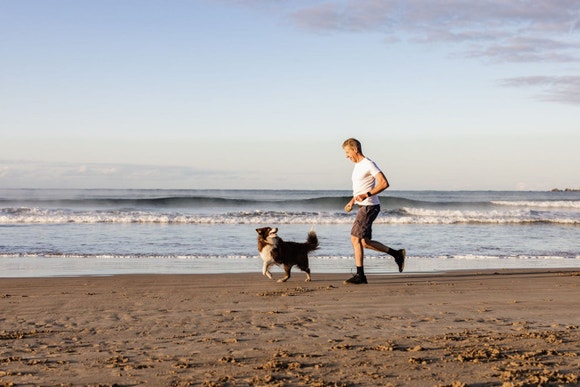 Man in white t-shirt running along beach with brown Border Collie