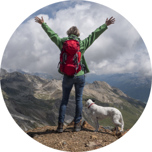 A white and black dog and a woman standing at the top of a mountain looking back at other mountains.